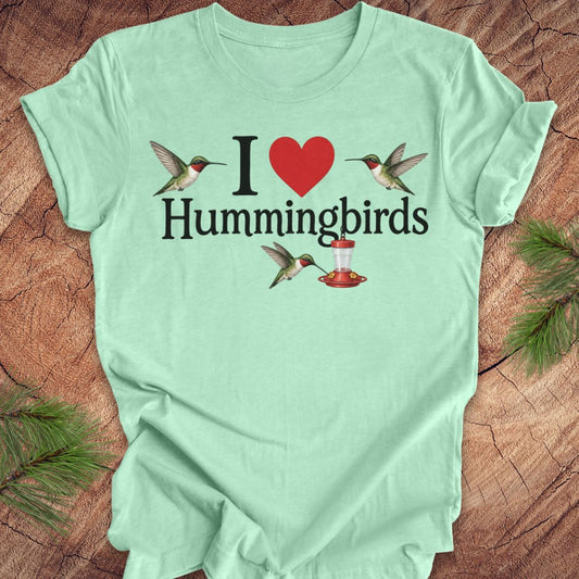 A mint green short-sleeve t-shirt laid flat on a rustic wood background with pine needles in the corners. The shirt features a graphic print that reads 'I [red heart] Hummingbirds' with illustrations of hummingbirds and a red-and-white hummingbird feeder.