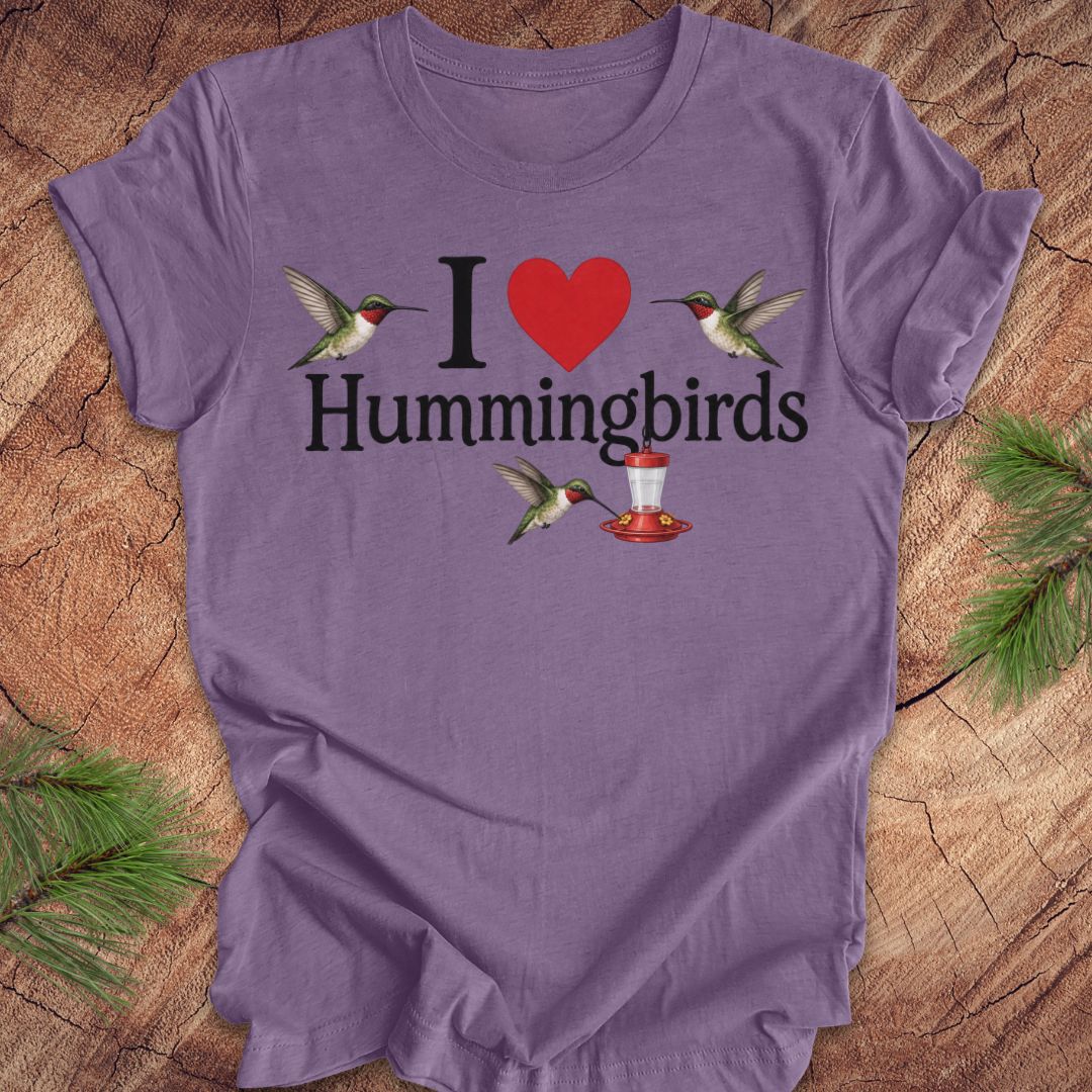 A purple short-sleeve t-shirt laid flat on a rustic wood surface with pine branches around it. The shirt features a graphic that reads 'I' followed by a red heart, then 'Hummingbirds' in black text, with two hummingbirds and a red-and-white hummingbird feeder illustrated around the text.