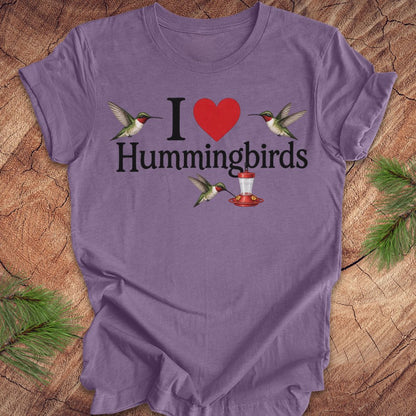 A purple short-sleeve t-shirt laid flat on a rustic wood surface with pine branches around it. The shirt features a graphic that reads 'I' followed by a red heart, then 'Hummingbirds' in black text, with two hummingbirds and a red-and-white hummingbird feeder illustrated around the text.