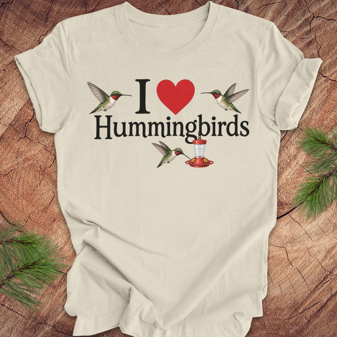 A beige t-shirt laid flat on a rustic wood background with pine branches in the corners. The shirt features a graphic print that reads 'I [red heart] Hummingbirds' with illustrations of hummingbirds and a hummingbird feeder. 
