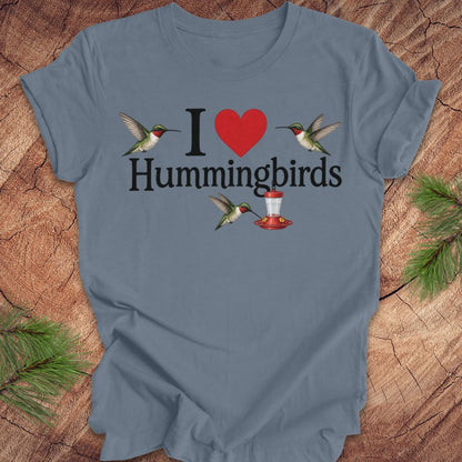 A slate blue crewneck t-shirt featuring a 'I [red heart] Hummingbirds' graphic with two hummingbirds and a red hummingbird feeder, laid flat on a rustic wood background with pine branches.