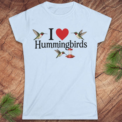 A light blue short-sleeve crewneck t-shirt laid flat on a rustic wood background with pine needles. The shirt features a graphic print that reads 'I' followed by a red heart, then 'Hummingbirds' in black text, with three hummingbird illustrations surrounding the text.