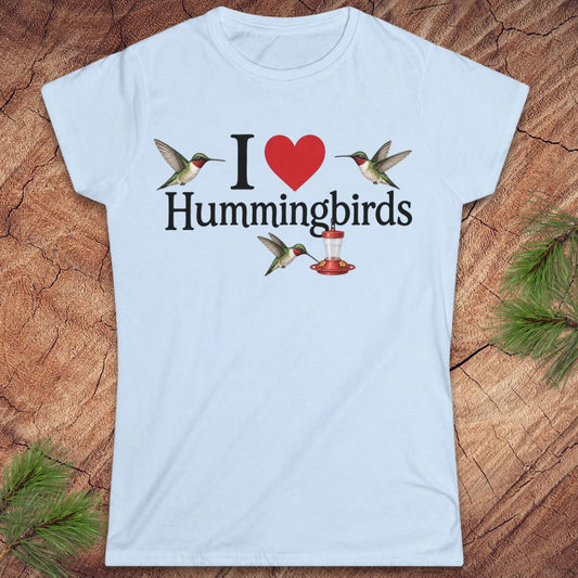 A light blue short-sleeve crewneck t-shirt laid flat on a rustic wood background with pine needles. The shirt features a graphic print that reads 'I' followed by a red heart, then 'Hummingbirds' in black text, with three hummingbird illustrations surrounding the text.