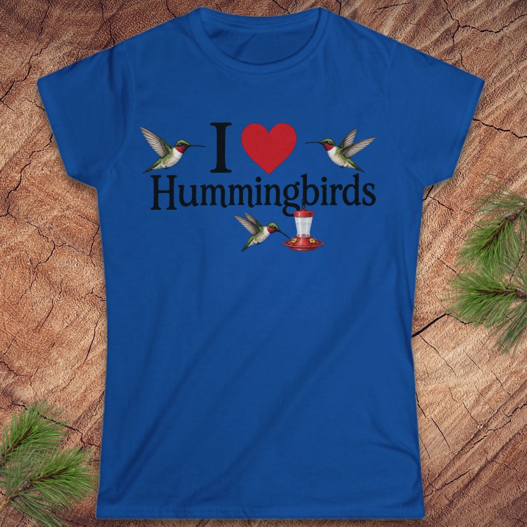 A royal blue short-sleeve crewneck t-shirt laid flat on a textured wood background with pine needles in the corners. The shirt features a graphic print that reads 'I [red heart] Hummingbirds' with illustrations of hummingbirds and a red hummingbird feeder.