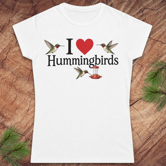 A white short-sleeve crewneck t-shirt featuring a graphic print that reads 'I [red heart] Hummingbirds' with two hummingbirds and a feeder, displayed flat on a rustic wood background with pine needles.
