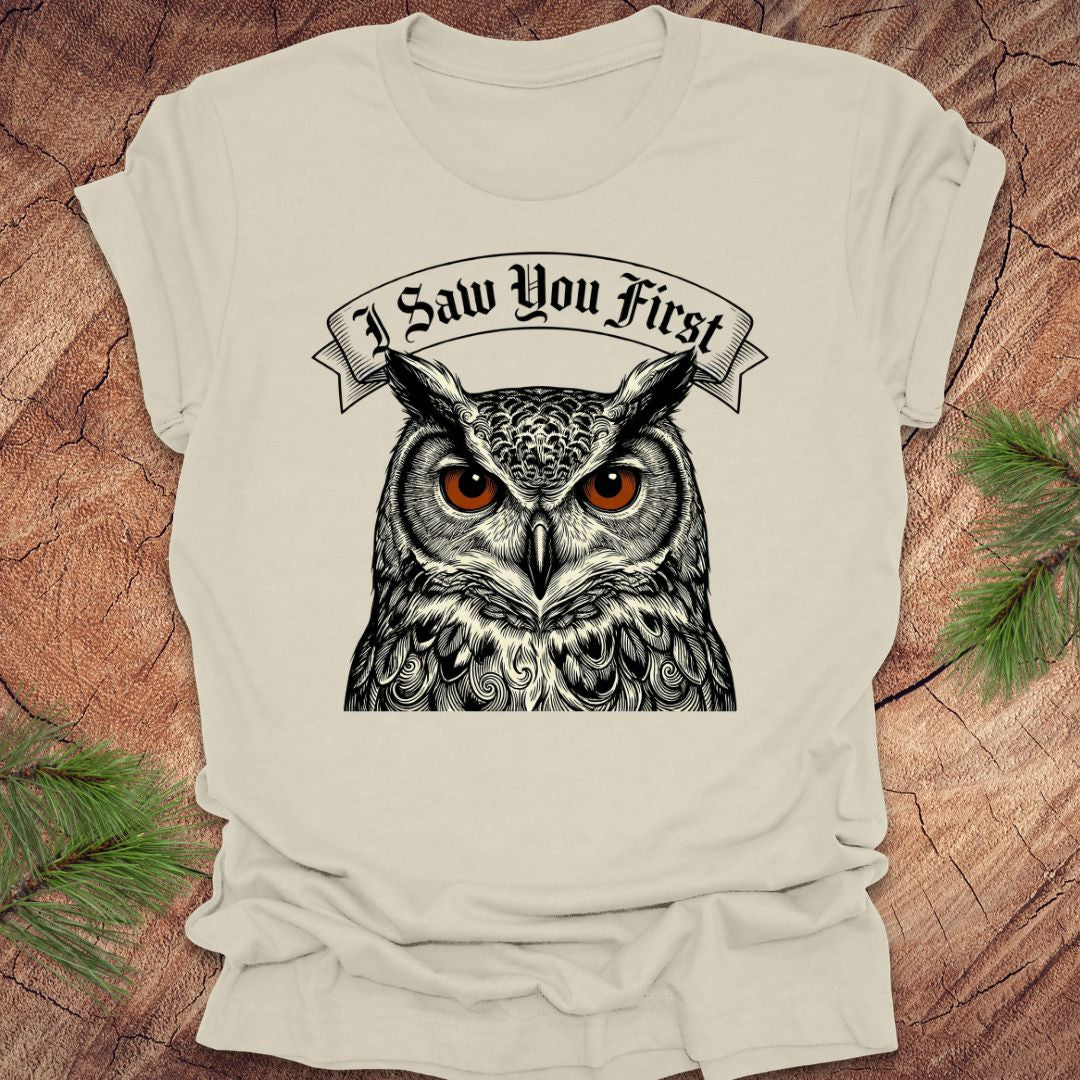 Beige t-shirt with an owl graphic and 'I Saw You First' text on a wooden surface.