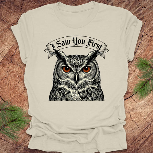 Beige t-shirt with an owl graphic and 'I Saw You First' text on a wooden surface.