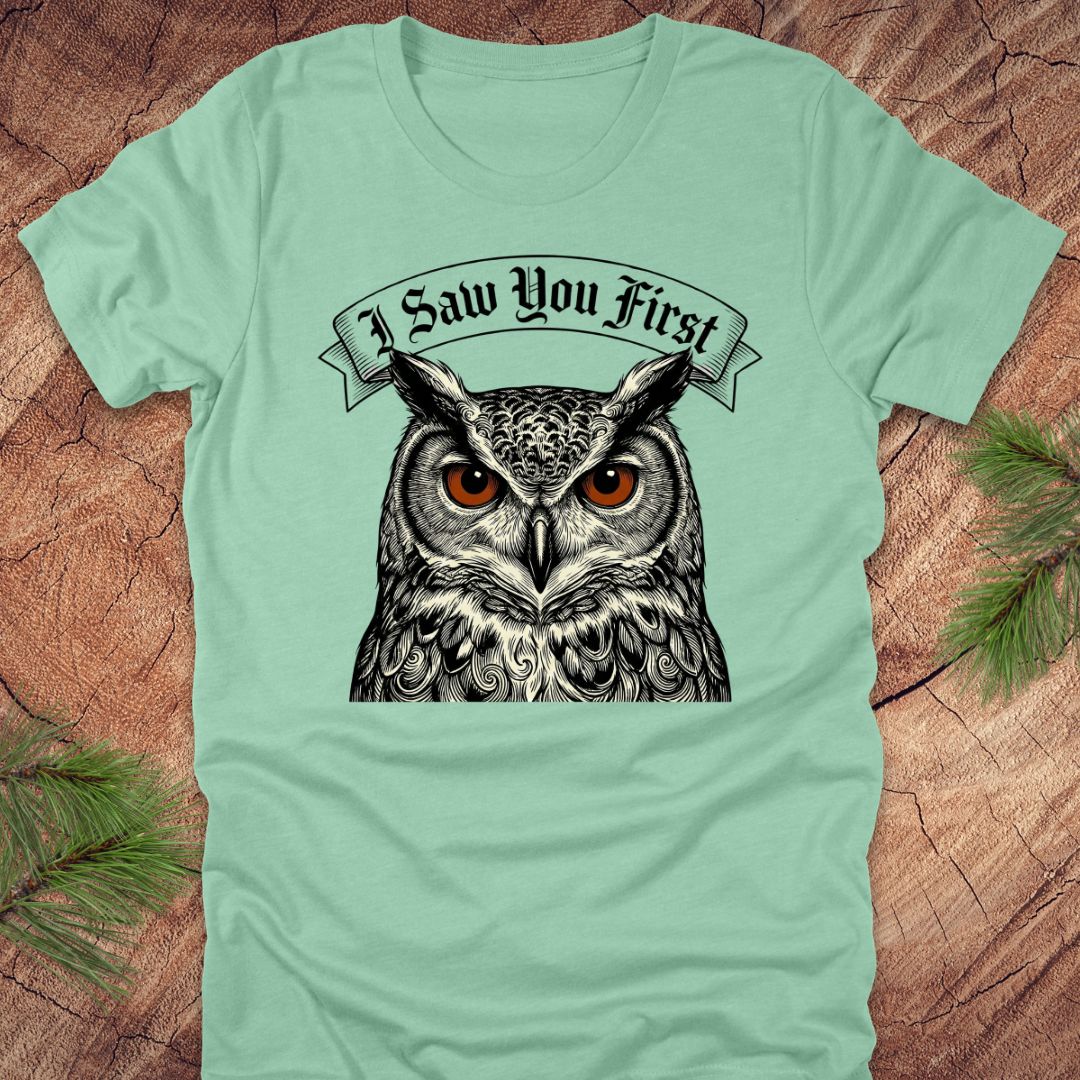Mint green t-shirt with an owl graphic and 'I Saw You First' text on a wooden surface.