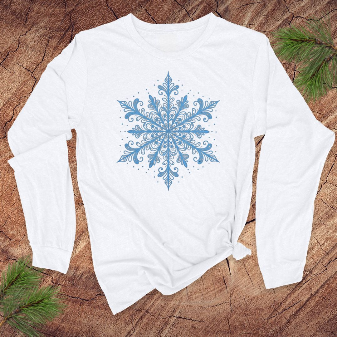 White long-sleeve shirt with an icy blue snowflake design on a wooden surface with pine branches.