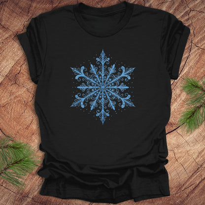 Black t-shirt with an icy blue snowflake design on a wooden surface with pine branches.