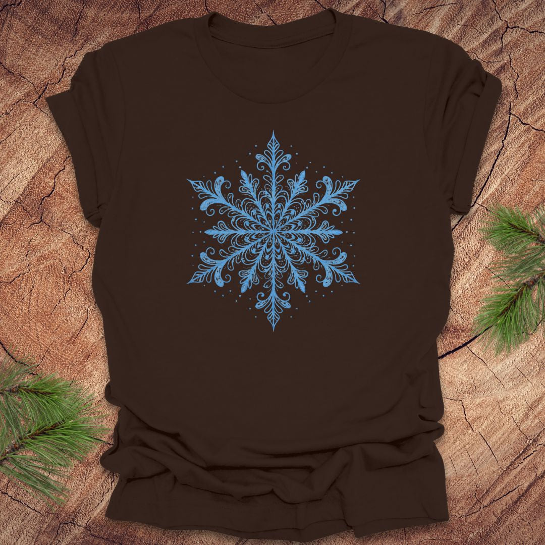 Brown t-shirt with an icy blue snowflake design on a wooden surface with pine branches.