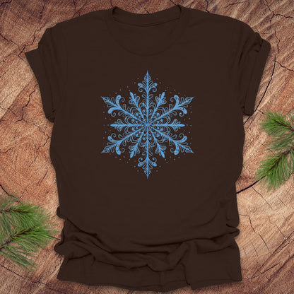 Brown t-shirt with an icy blue snowflake design on a wooden surface with pine branches.