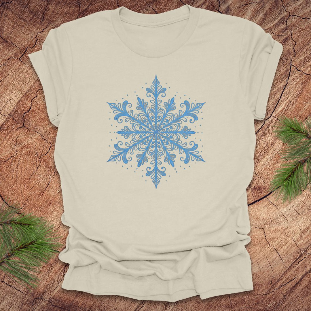 Beige t-shirt with an icy blue snowflake design on a wooden surface with pine branches.
