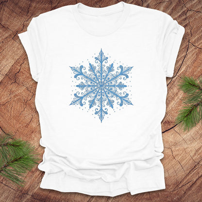 White t-shirt with an icy blue snowflake design on a wooden surface with pine branches.