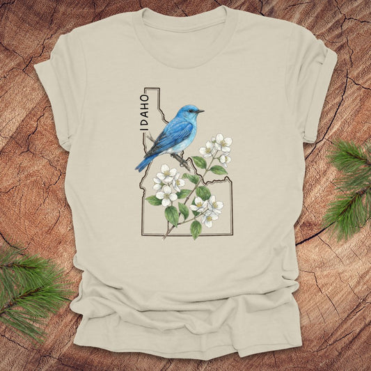 Beige t-shirt with Idaho state outline and state bird, mountain bluebird design with flowers on a wooden surface