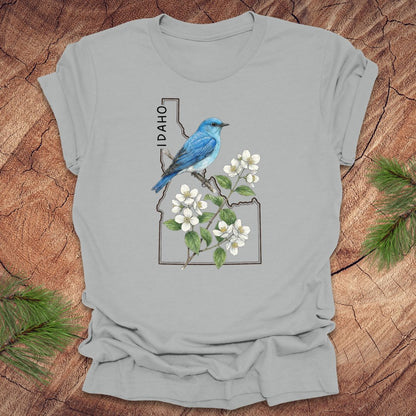 Silver t-shirt with Idaho state outline and state bird, mountain bluebird design with flowers on a wooden surface