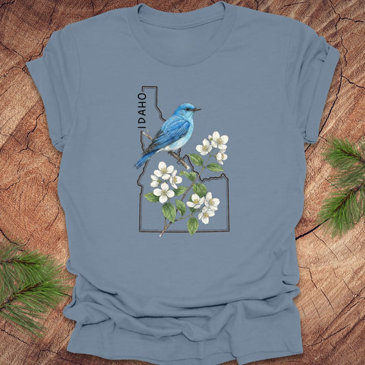 Steel blue t-shirt with Idaho state outline and state bird, mountain bluebird design with flowers on a wooden surface