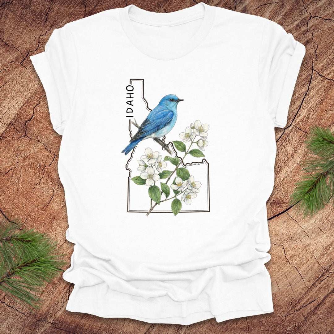 White t-shirt with Idaho state outline and state bird, mountain bluebird design with flowers on a wooden surface