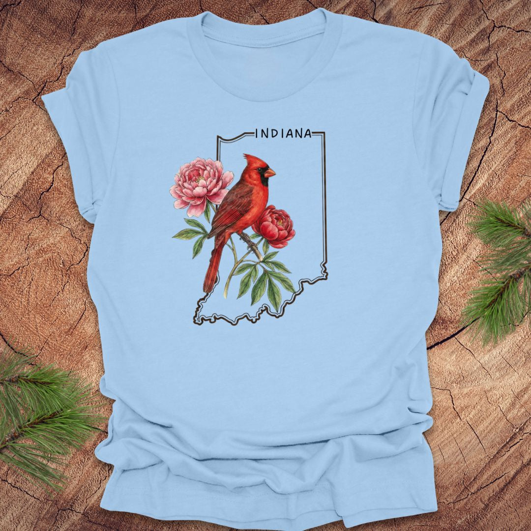 Baby blue t-shirt with a red cardinal and peony design on an outline of Indiana, placed on a wooden surface.