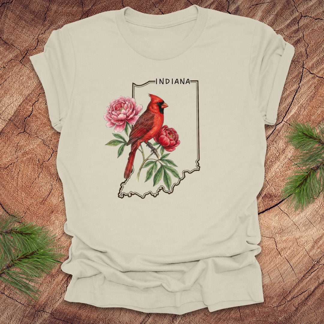 Beige t-shirt with a red cardinal and peony design on an outline of Indiana, placed on a wooden surface.