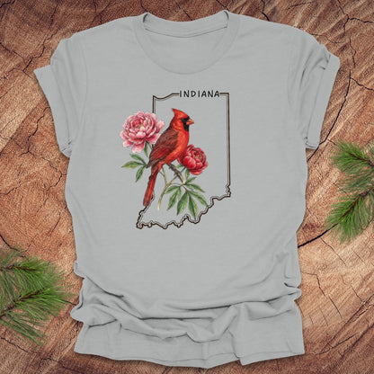 Silver t-shirt with a red cardinal and peony design on an outline of Indiana, placed on a wooden surface.