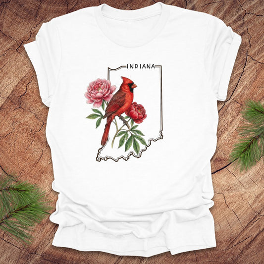 White t-shirt with a red cardinal and peony design on an outline of Indiana, placed on a wooden surface.