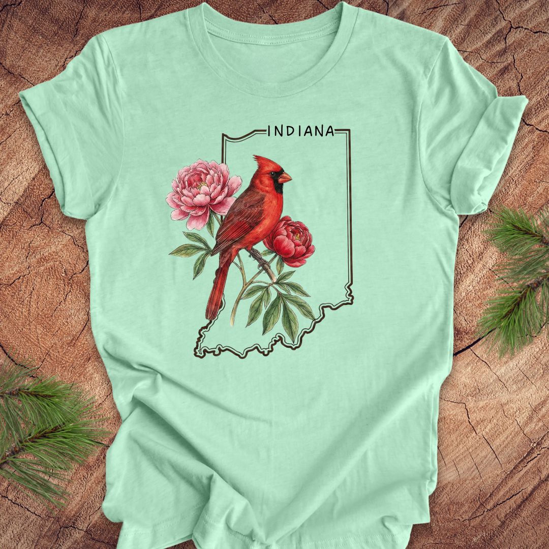 Mint green t-shirt with a red cardinal and peony design on an outline of Indiana, placed on a wooden surface.
