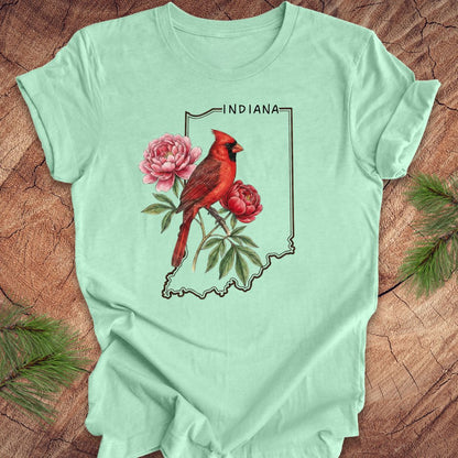 Mint green t-shirt with a red cardinal and peony design on an outline of Indiana, placed on a wooden surface.
