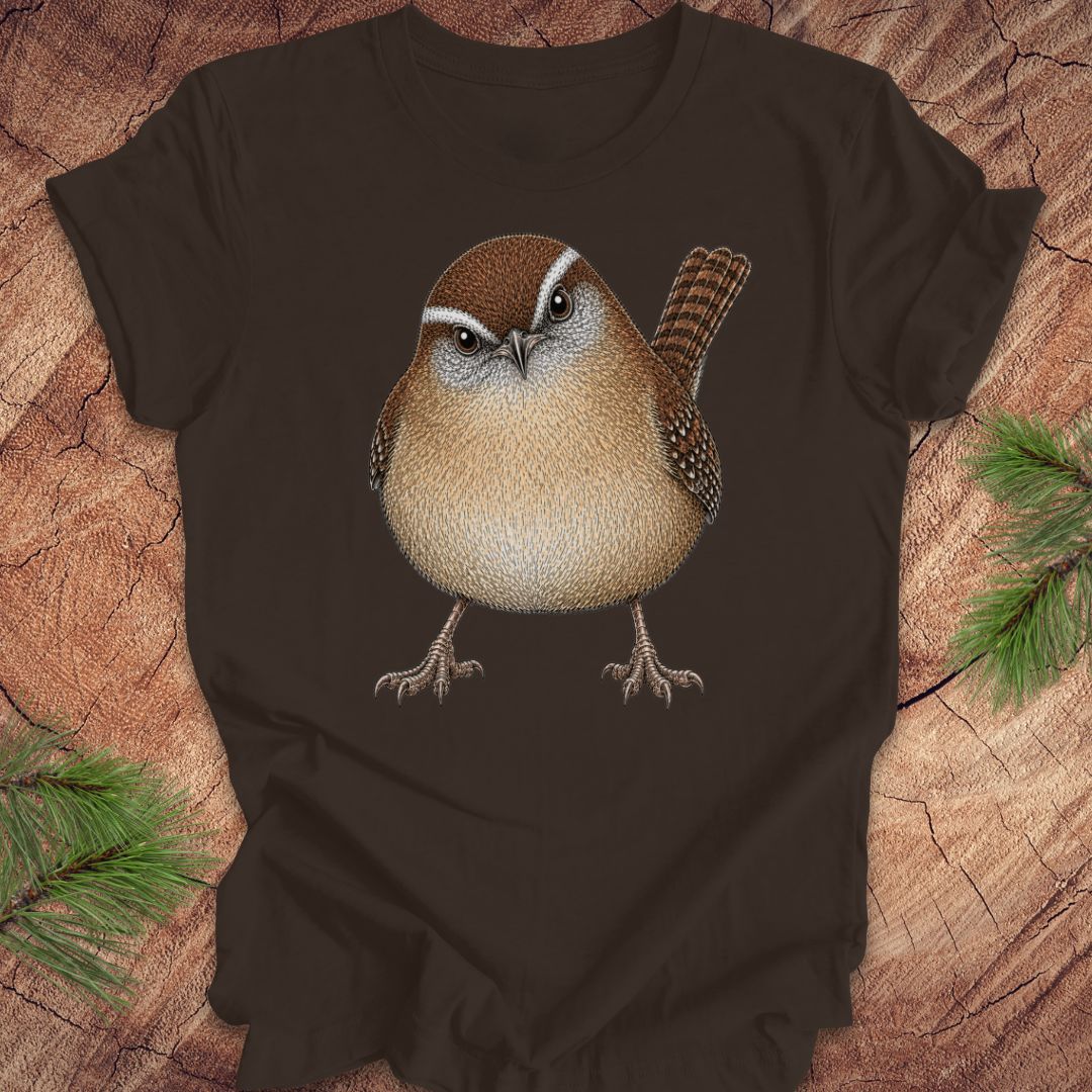 Brown t-shirt with a carolina wren design on a wooden surface with pine branches.