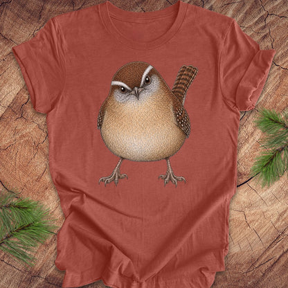 Rust colored t-shirt with a carolina wren design on a wooden surface with pine branches.