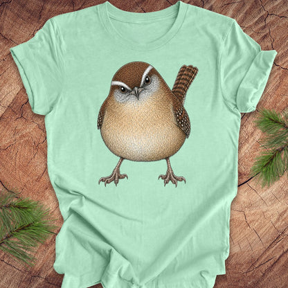 Mint green t-shirt with a carolina wren design on a wooden surface with pine branches.