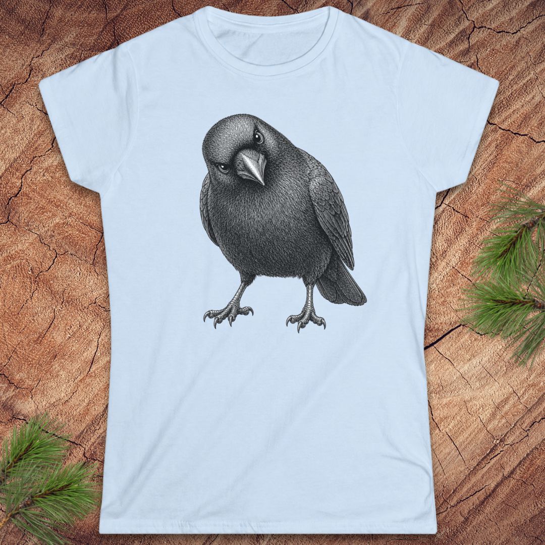 Light blue women's t-shirt with a detailed pencil-style American Crow tilting its head and glaring, laid flat on a rustic wood background with pine branches.