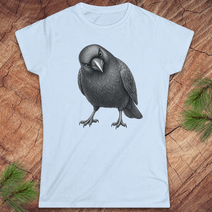 Light blue women's t-shirt with a detailed pencil-style American Crow tilting its head and glaring, laid flat on a rustic wood background with pine branches.