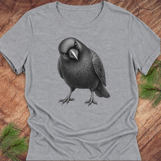 Athletic heather gray women's relaxed t-shirt with a pencil-style American Crow tilting its head and giving a judgy stare, laid flat on a rustic wood background with pine branches.