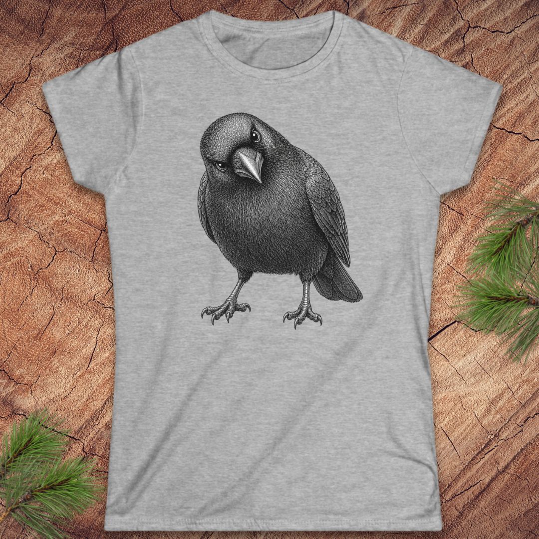 Sport grey women's t-shirt with a detailed pencil-style American Crow tilting its head and glaring, laid flat on a rustic wood background with pine branches.