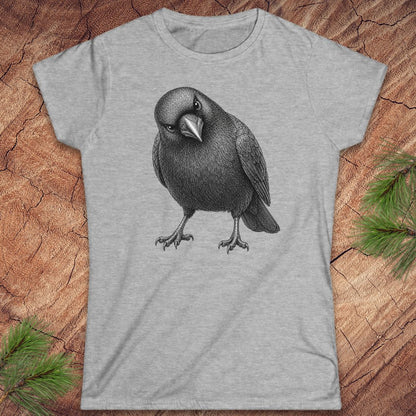 Sport grey women's t-shirt with a detailed pencil-style American Crow tilting its head and glaring, laid flat on a rustic wood background with pine branches.