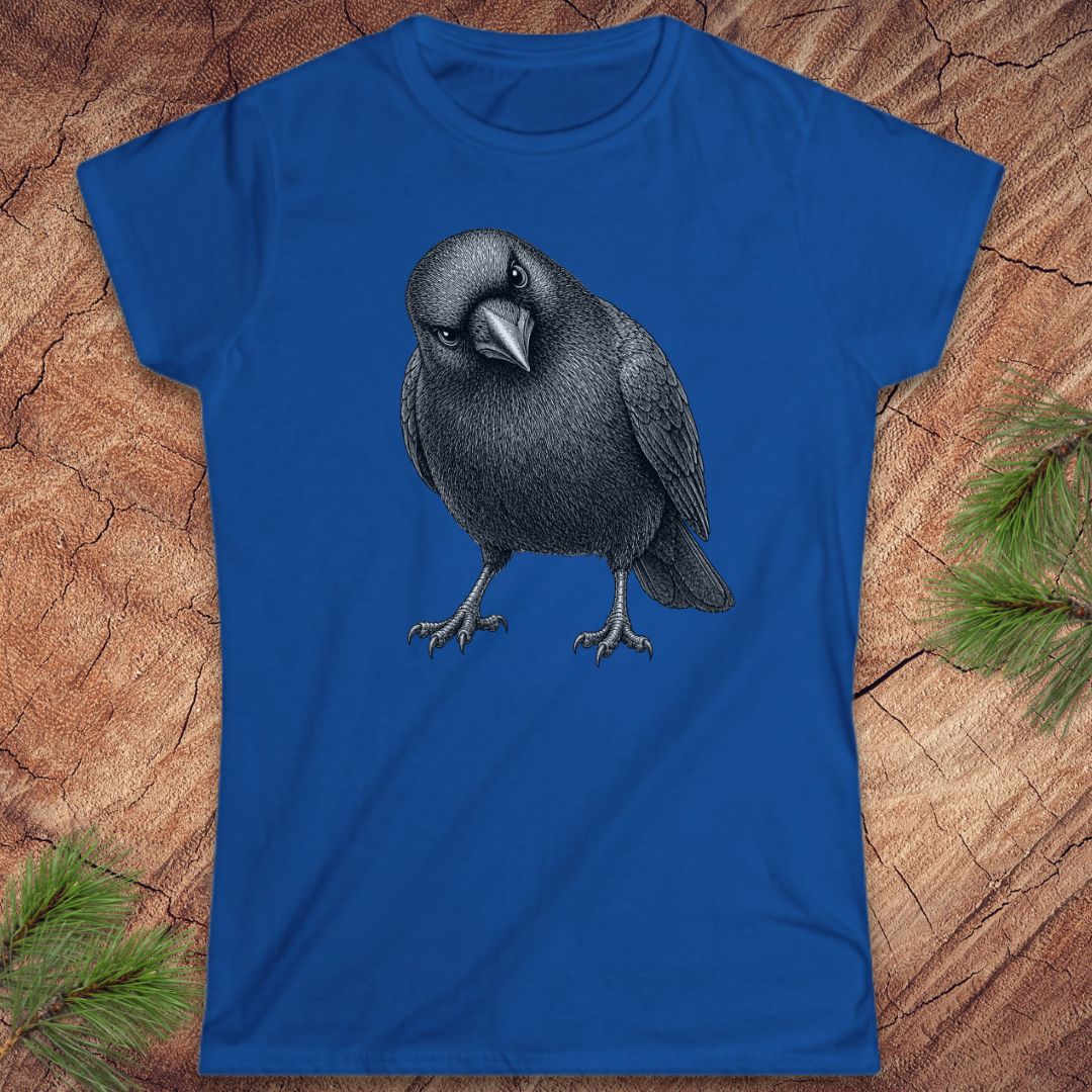 Royal blue women's t-shirt with a detailed pencil-style American Crow tilting its head and glaring, laid flat on a rustic wood background with pine branches.