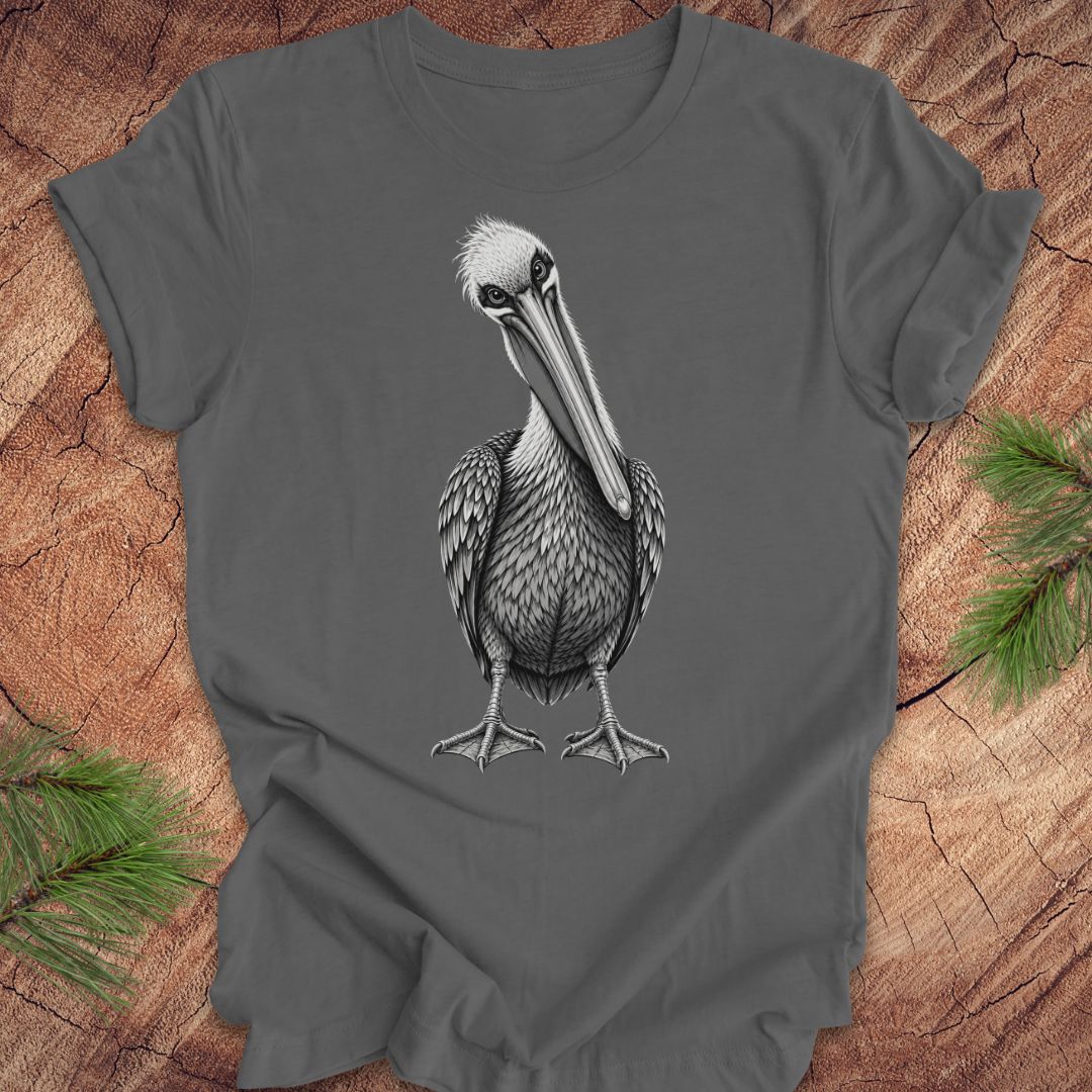 A dark gray t-shirt featuring a detailed black and white graphic of a pelican bird, laid flat on a rustic wood background with pine needles in the corners.