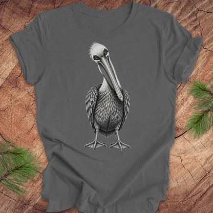 A dark gray t-shirt featuring a detailed black and white graphic of a pelican bird, laid flat on a rustic wood background with pine needles in the corners.