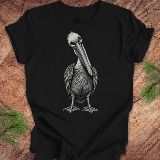 A black short-sleeve crewneck t-shirt featuring a detailed black and white illustration of a brown pelican on the front, displayed flat on a rustic wood background with pine needles in the corners.