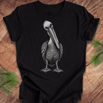 A black short-sleeve crewneck t-shirt featuring a detailed black and white illustration of a pelican on the front, displayed flat on a rustic wood background with pine branches in the corners.