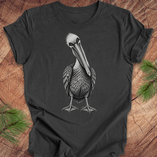 A dark charcoal gray t-shirt featuring a detailed black and white graphic of a standing pelican, laid flat on a rustic wood background with pine needles in the corners.