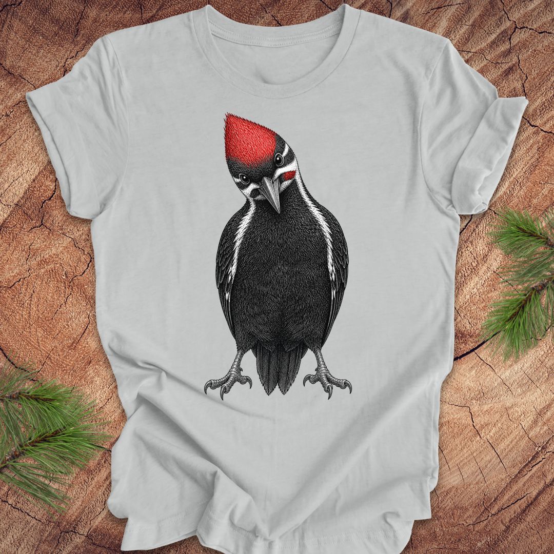 Gray t-shirt with a woodpecker design on a wooden surface with pine branches.