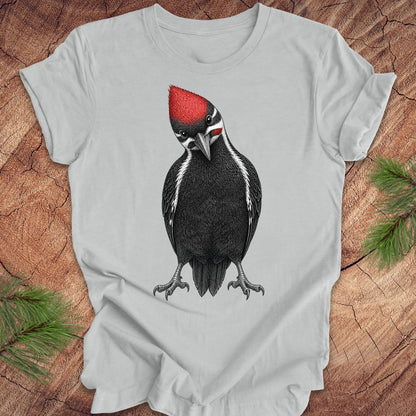 Gray t-shirt with a woodpecker design on a wooden surface with pine branches.