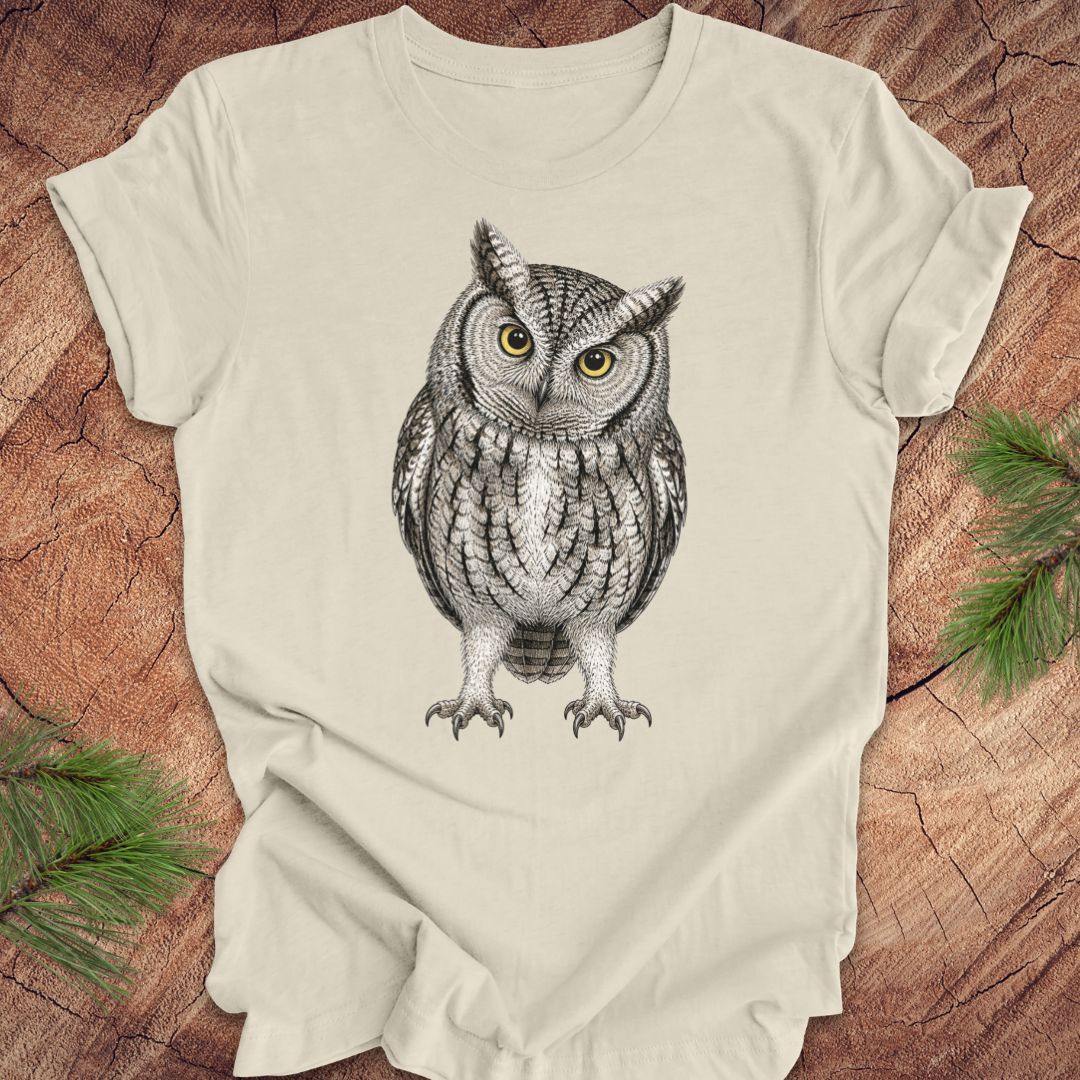 Beige t-shirt with an owl design on a wooden surface with green leaves.