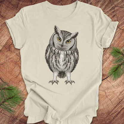 Beige t-shirt with an owl design on a wooden surface with green leaves.