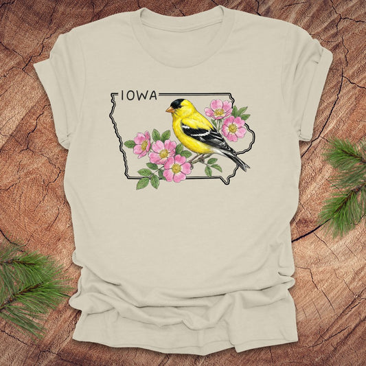 Beige t-shirt with the outline of the state of Iowa with state bird and flower, goldfinch and rose, on a wood background.