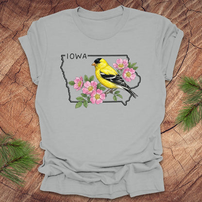 Silver t-shirt with the outline of the state of Iowa with state bird and flower, goldfinch and rose, on a wood background.