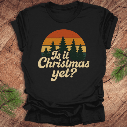 Black t-shirt with 'Is it Christmas yet?' design on a wooden surface with pine branches.