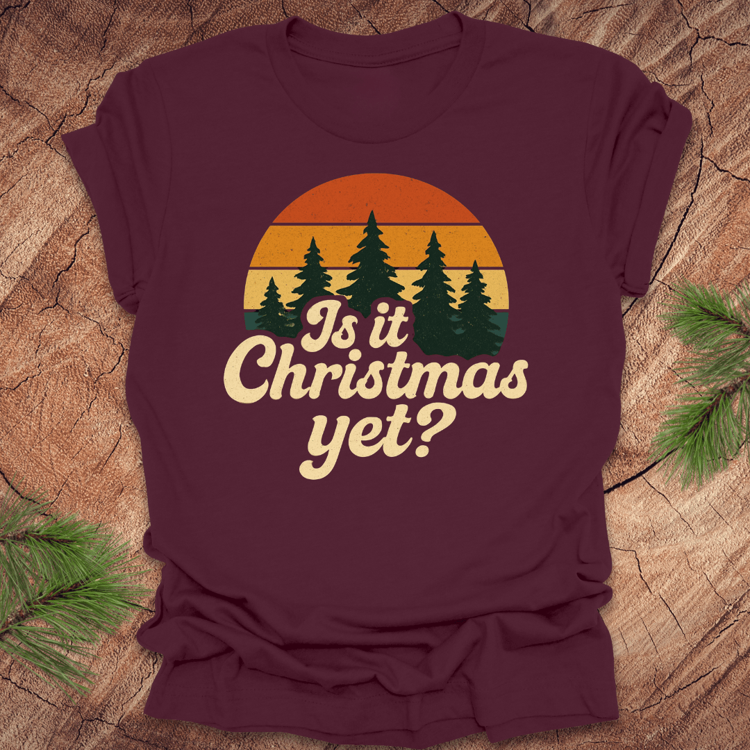 Maroon t-shirt with 'Is it Christmas yet?' graphic on a wooden surface with pine branches.